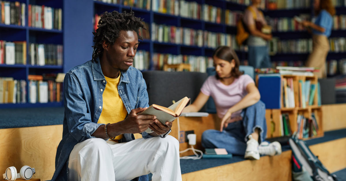 Portrait of young black man reading book in library lounge with students in background, copy space