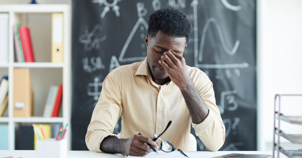 Tired young teacher sitting by desk in classroom and touching nose bridge after several lessons