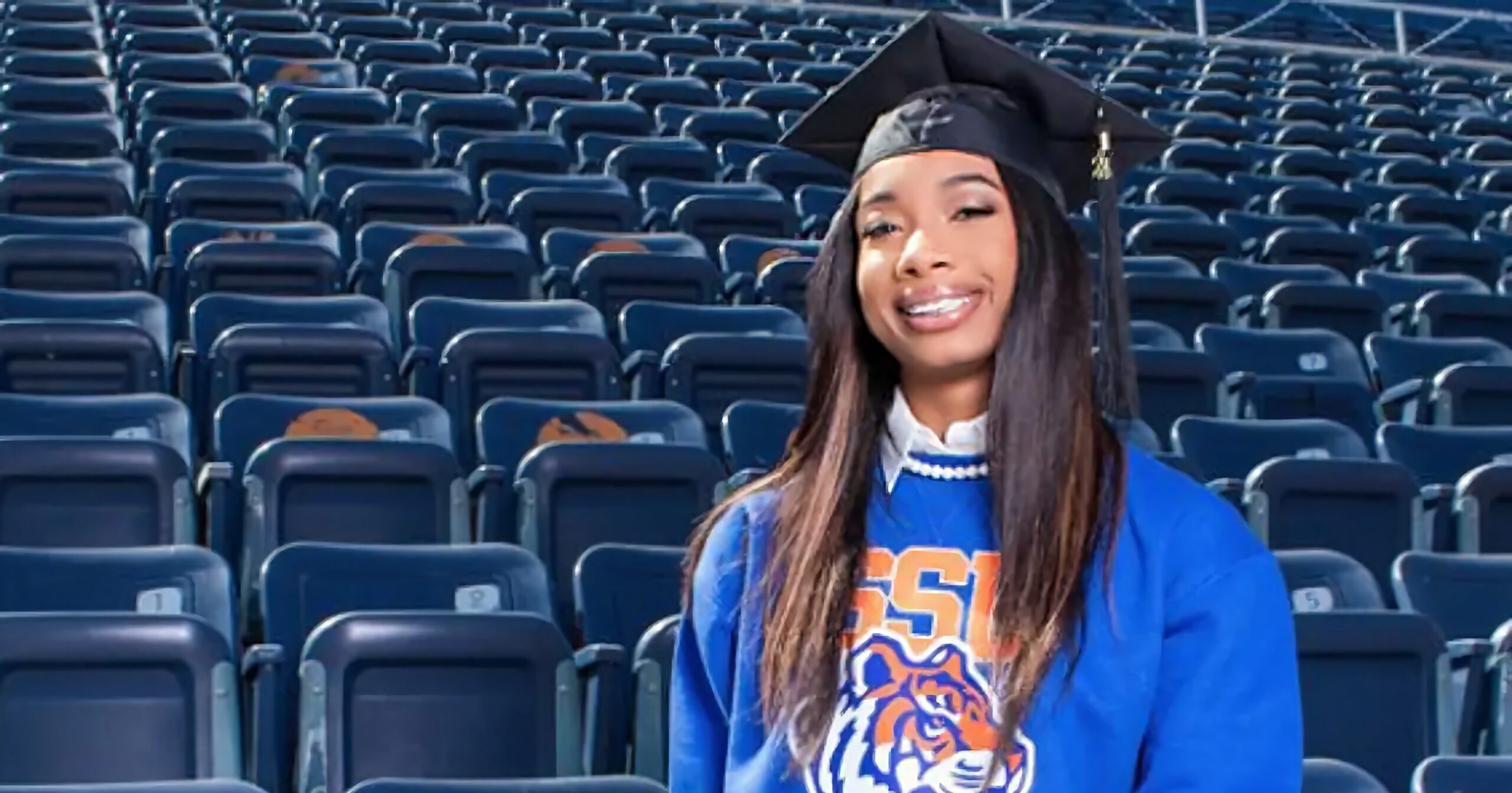 NeeAli Scott wearing graduation cap and college sweatshirt standing in front of bleacher seats.
