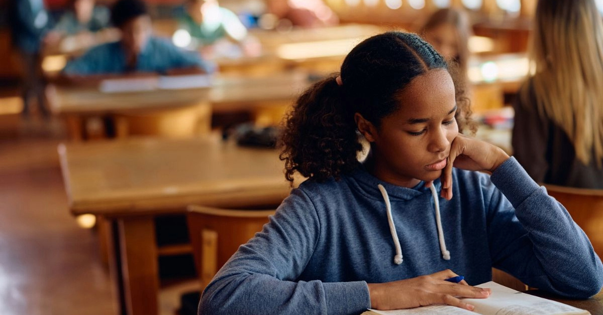 A young Black female student reads a book in a classroom.